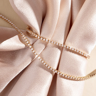 A gold tennis bracelet and necklace set laid across a light pink velvet fabric on a beige background.