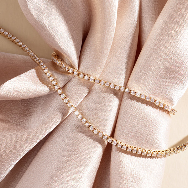 A gold tennis bracelet and necklace set laid across a light pink velvet fabric on a beige background.