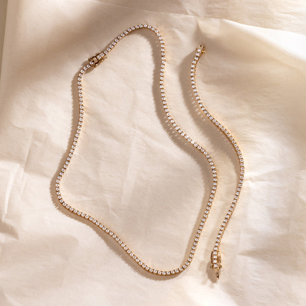 A gold tennis bracelet and necklace laid side by side on a textured white background.