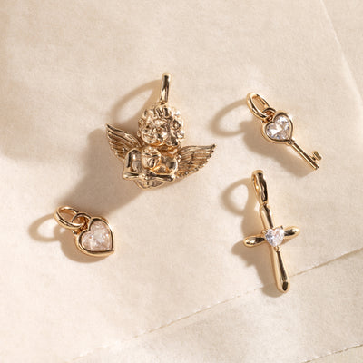 Four gold charms including a cherub, key, heart, and cross on a beige fabric background