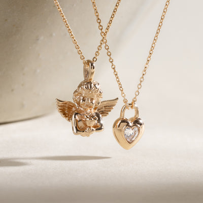 Two gold pendant necklaces with a cherub and a padlock heart charm on a neutral background