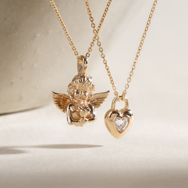 Two gold pendant necklaces with a cherub and a padlock heart charm on a neutral background
