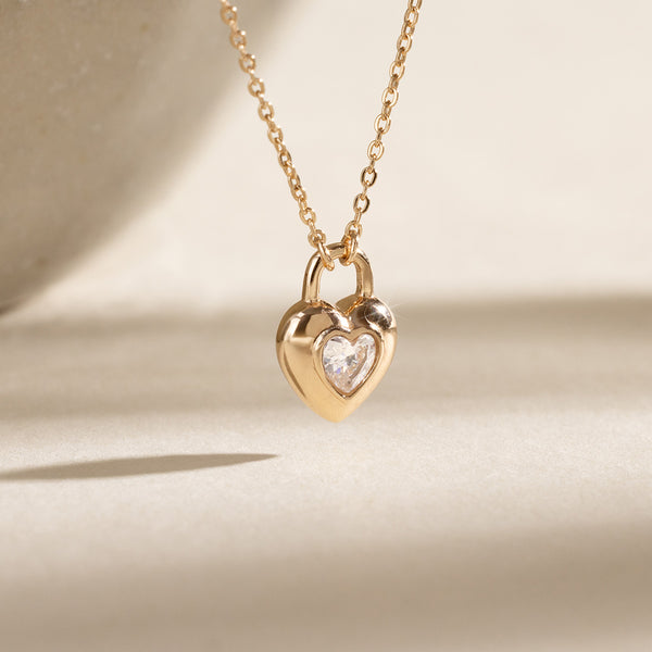 A gold chain pendant necklace with a heart padlock shaped charm, featuring a heart-shaped clear stone in the center, set against a warm white backdrop.