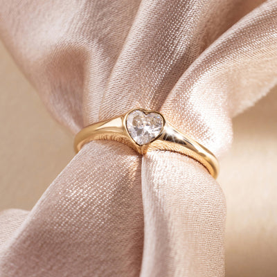 Gold signet ring with a heart-shaped clear stone on a soft pink fabric background