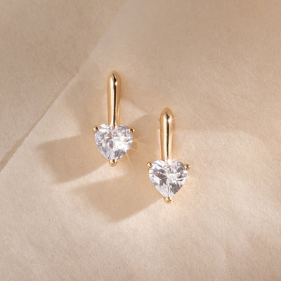 A pair of gold drop stud earrings with heart-shaped prong set clear stones, set against a textured cream background.