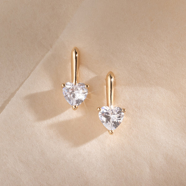 A pair of gold drop stud earrings with heart-shaped prong set clear stones, set against a textured cream background.