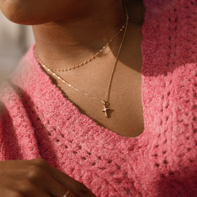 Person wearing a pink knitted garment and gold necklace with a cross pendant featuring a heart shaped clear stone in the center.