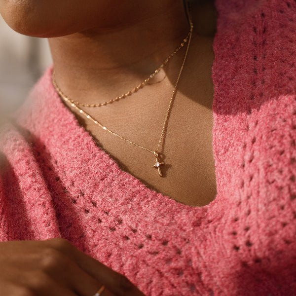 Person wearing a pink knitted garment and gold necklace with a cross pendant featuring a heart shaped clear stone in the center.