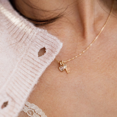 Close-up lifestyle image of a person wearing a gold dainty chain necklace with a clear stone heart and a gold heart key shaped pendant.