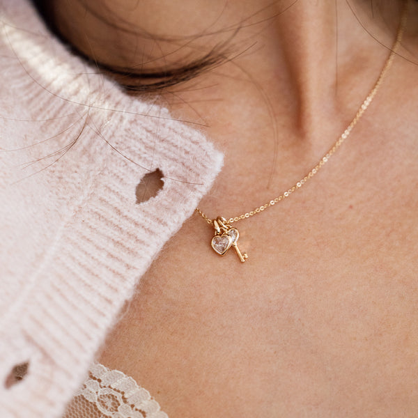 Close-up lifestyle image of a person wearing a gold dainty chain necklace with a clear stone heart and a gold heart key shaped pendant.