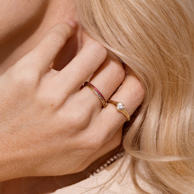 A close up lifestyle image of a hand along a neck with blonde hair, showcasing two gold stacked rings with different colored stones, and a gold signet ring with a heart shaped clear stone in the center.