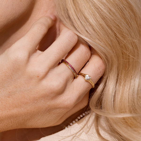 A close up lifestyle image of a hand along a neck with blonde hair, showcasing two gold stacked rings with different colored stones, and a gold signet ring with a heart shaped clear stone in the center.