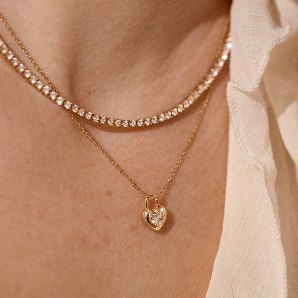 A close up lifestyle photo of a person wearing a gold tennis necklace with clear stones, and a dainty gold chain necklace with a gold heart padlock charm featuring a heart-shaped clear stone in the center.