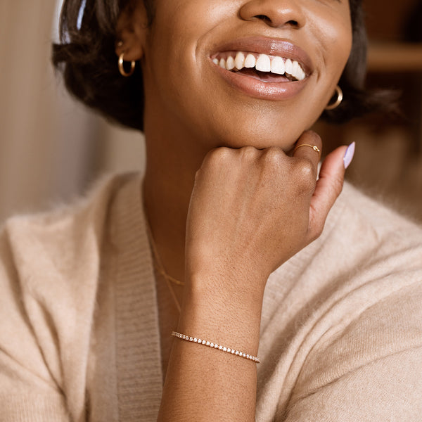 A lifestyle image of a woman wearing a beige sweater with a gold tennis bracelet on her wrist, accented by gold hoops and a gold ring.