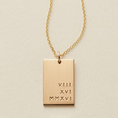A gold rectangular pendant necklace engraved with the Roman numerals 'VIII XVI MMXVI' on a textured cream background.