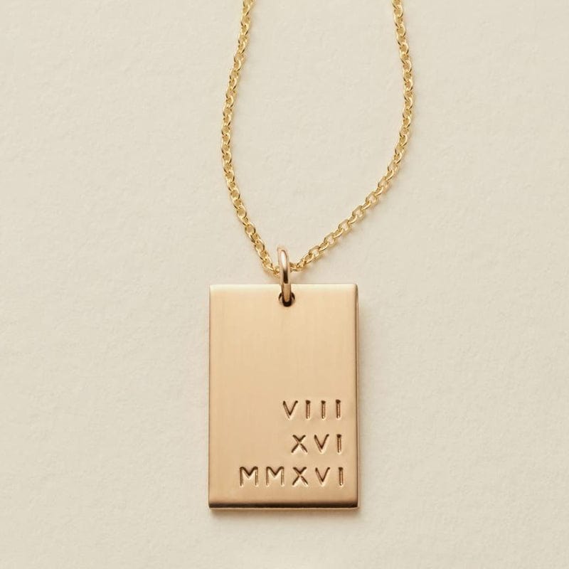A gold rectangular pendant necklace engraved with the Roman numerals 'VIII XVI MMXVI' on a textured cream background.