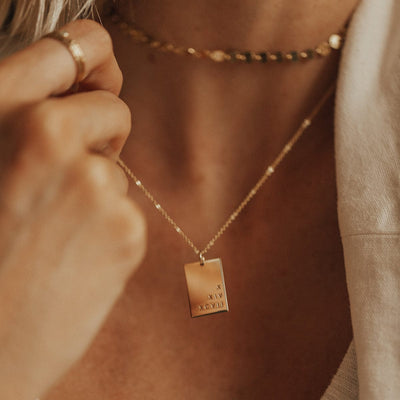 A close-up of a person's neck wearing a delicate gold pendant chain necklace with a flat rectangle charm, engraved with roman numerals.