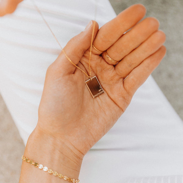 A person's hand holding a delicate gold rectangle pendant necklace, engraved with the words 'I AM ENOUGH' in the bottom right corner.
