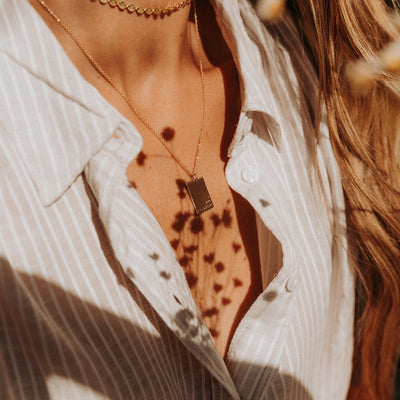 A close-up of a person's chest wearing a beige and white striped button up shirt paired with a delicate gold rectangle pendant necklace. There is direct sunlight on them, with shadows of small flowers.