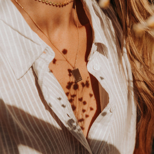 A close-up of a person's chest wearing a beige and white striped button up shirt paired with a delicate gold rectangle pendant necklace. There is direct sunlight on them, with shadows of small flowers.