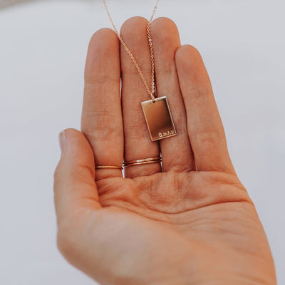 A person's hand holding a gold rectangle pendant necklace engraved with the phrase 'G>^v' in the bottom right corner, shown on a solid white background.
