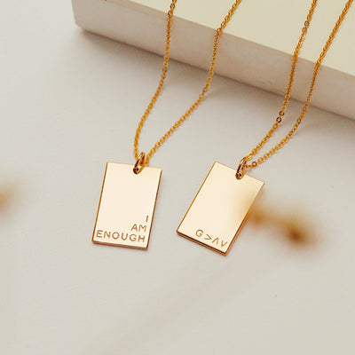 Two delicate gold pendant chain necklaces with flat rectangular charms. One is engraved with the phrase 'I AM ENOUGH' in three rows in the bottom right corner, and the other has 'G>^V' engraved in the bottom right corner.