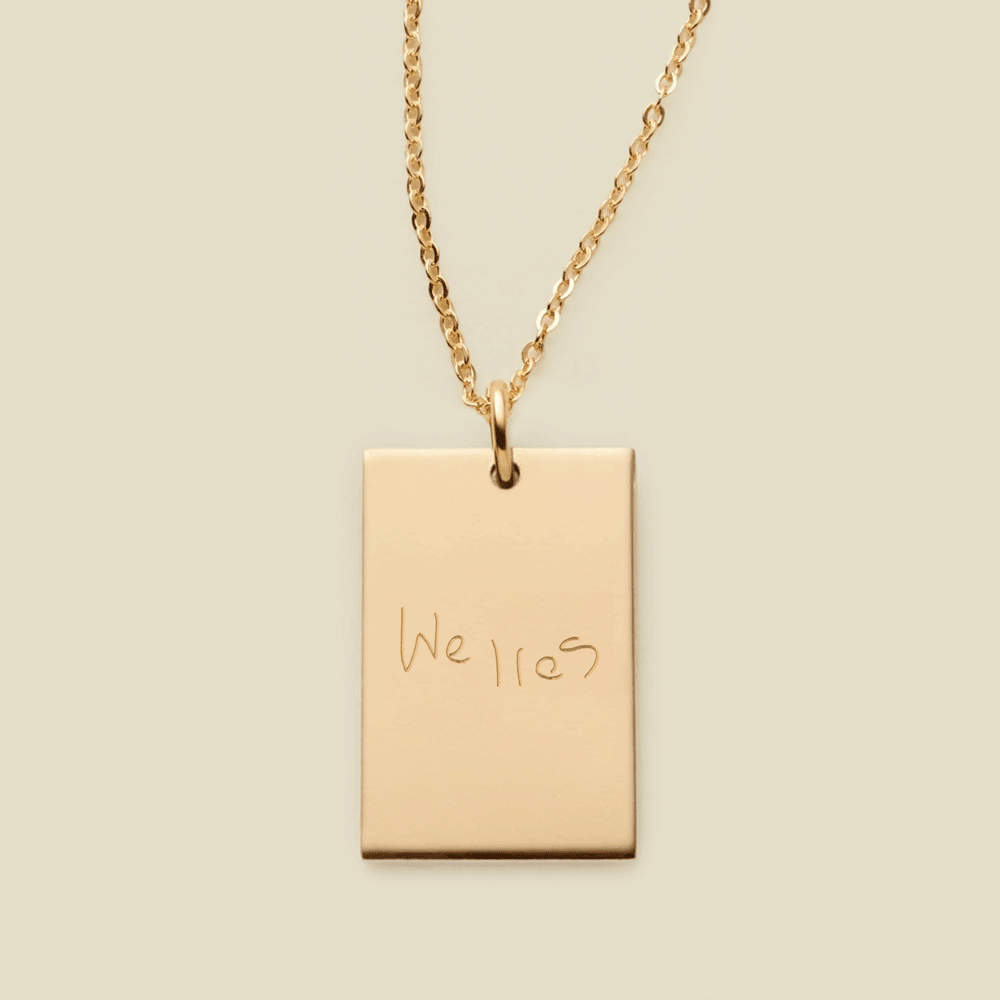 A dainty gold chain necklace with a rectangular gold pendant engraved with handwritten script.