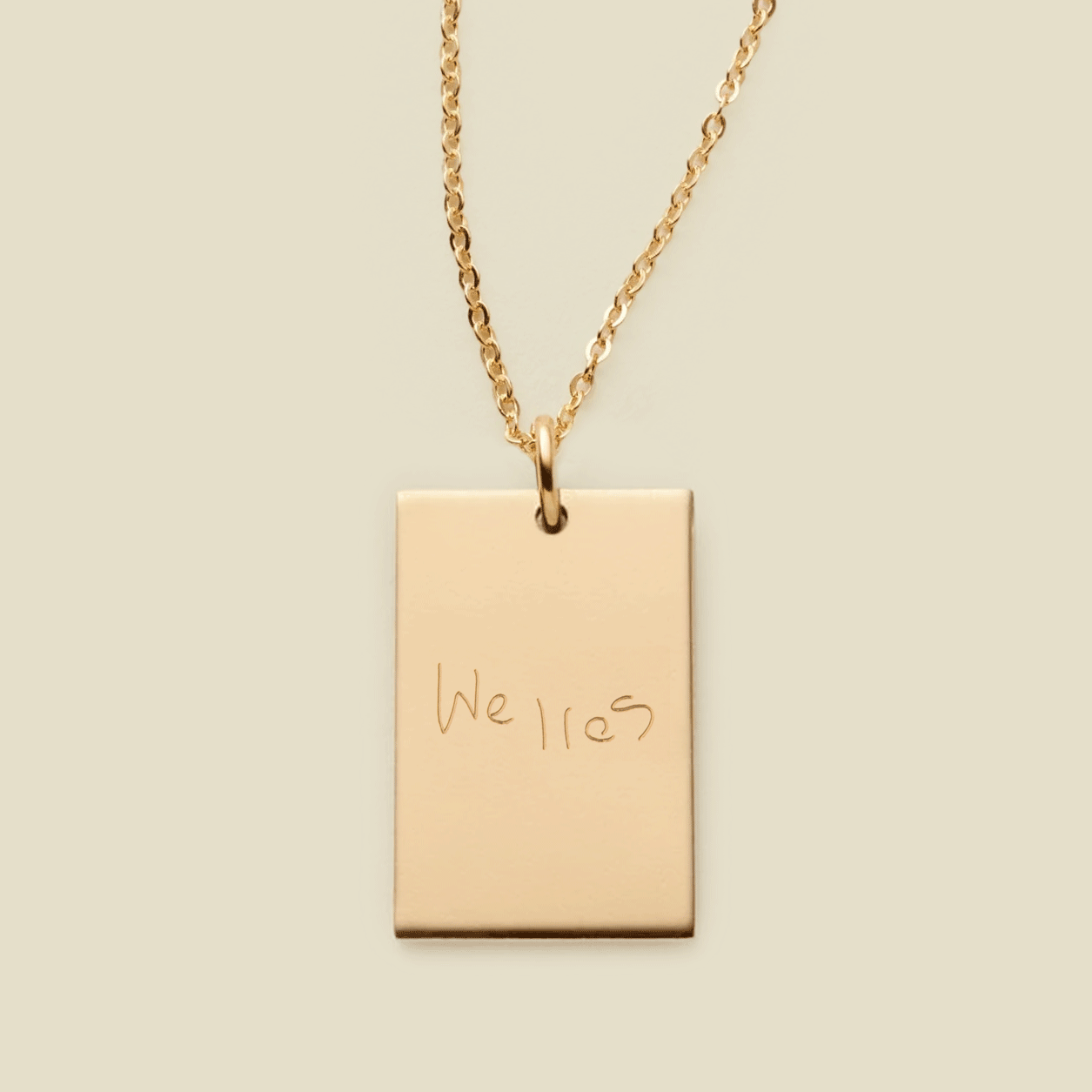 A dainty gold chain necklace with a rectangular gold pendant engraved with handwritten script.
