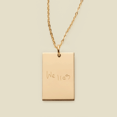 A dainty gold chain necklace with a rectangular gold pendant engraved with handwritten script.
