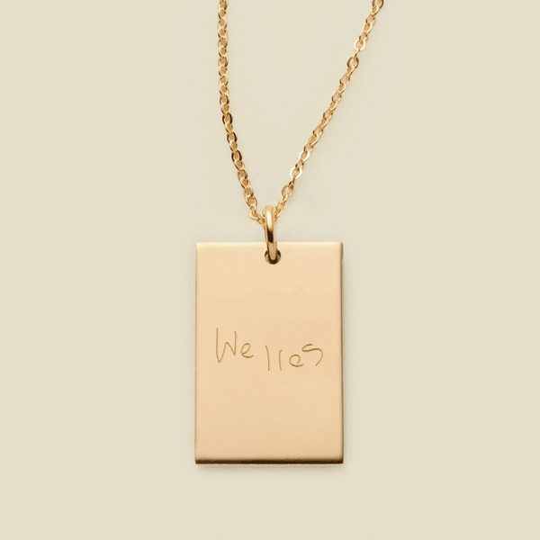 A dainty gold chain necklace with a rectangular gold pendant engraved with handwritten script.