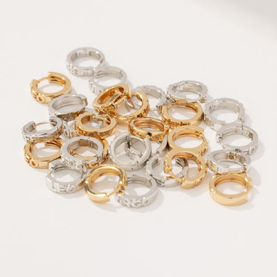 Multiple gold and silver huggie hoop earrings scattered on a solid white surface. Each earring has the name of a zodiac sign in raised letters across the front.