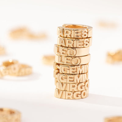 A stack of four pairs of gold huggie hoop earrings on a white surface. Each of the earrings has the name of a zodiac sign in raised letters across the front.