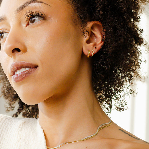 A person's ear wearing two gold hoop earrings, one with clear round gemstones in a curving pattern and the other with the word 'LEO' in raised letters along the front.