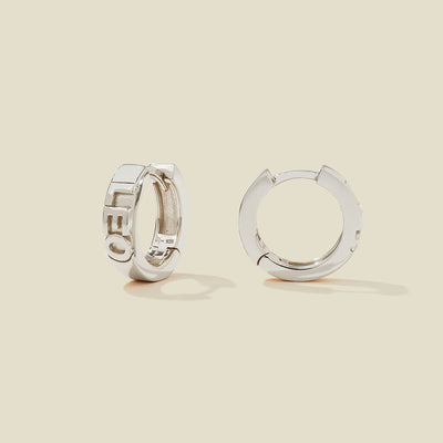 A pair of silver huggie hoop earrings on a light beige background. One earring is shown from the side, displaying a polished outer surface, while the other is shown from the front, showcasing the word 'LEO' in raised letters.