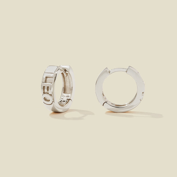 A pair of silver huggie hoop earrings on a light beige background. One earring is shown from the side, displaying a polished outer surface, while the other is shown from the front, showcasing the word 'LEO' in raised letters.