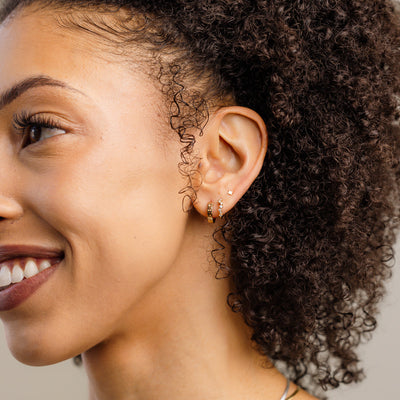 A close-up of a person's ear wearing two gold hoop earrings, one with clear round gemstones in a curving pattern and one with the word 'LIBRA' in raised letters.