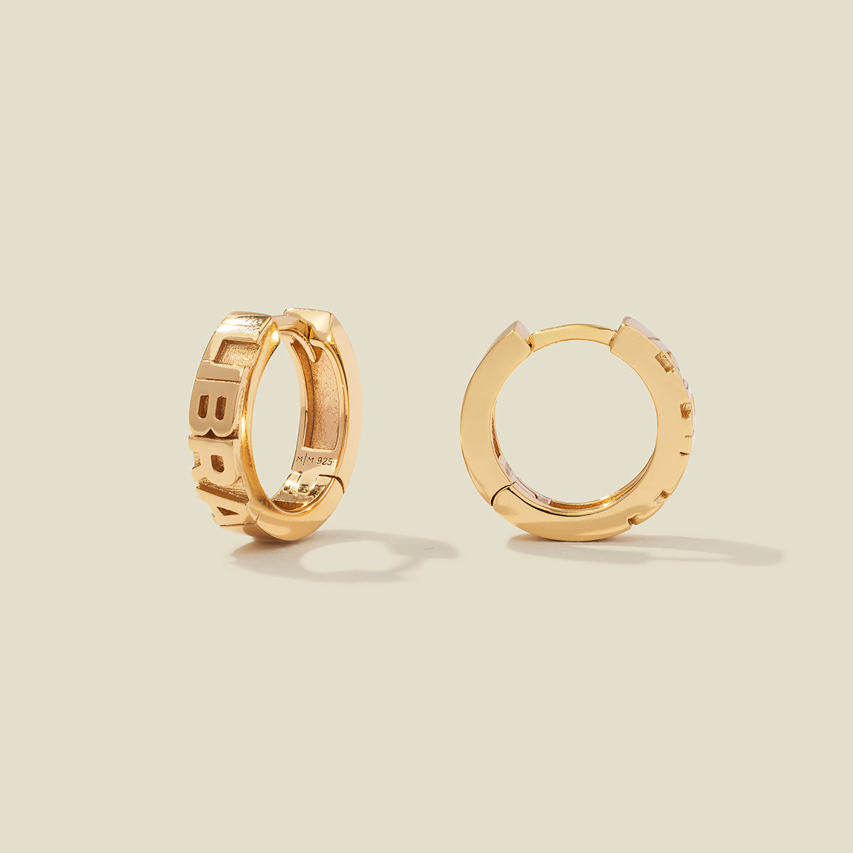 A pair of gold huggie hoop earrings on a light beige background. One earring is shown from the side, displaying a polished outer surface, while the other is shown from the front, showcasing the word 'LIBRA' in raised letters.
