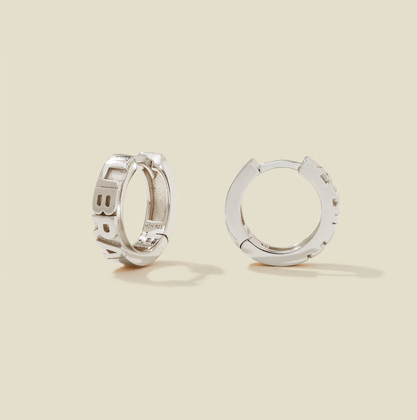 A pair of silver huggie hoop earrings on a light beige background. One earring is shown from the side, displaying a polished outer surface, while the other is shown from the front, showcasing the word 'LIBRA' in raised letters.