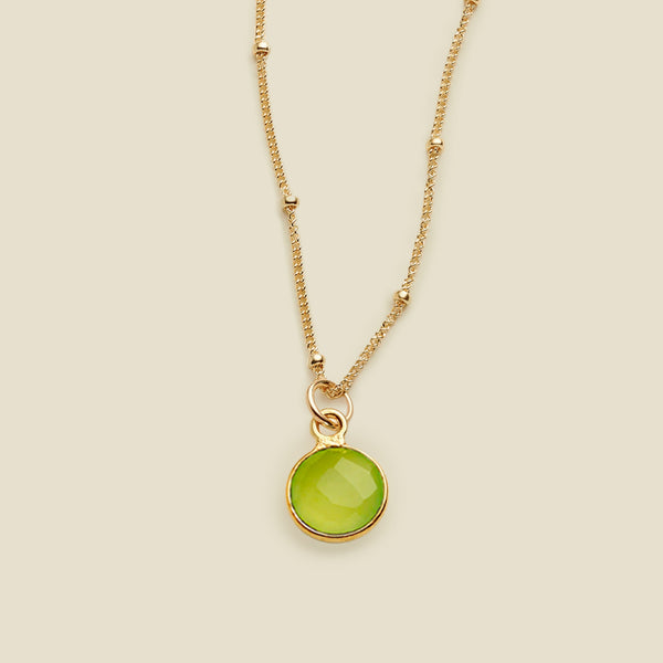 A gold chain pendant necklace with stationary gold beads, with a lime green gemstone charm, set against a solid beige background.
