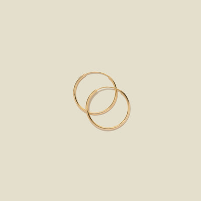 A pair of thin gold hoop earrings laying on top of one another, shown on a solid light beige background.