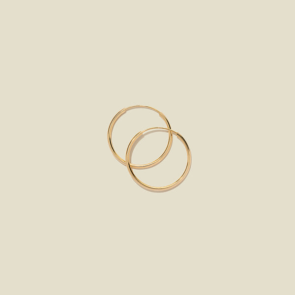 A pair of thin gold hoop earrings laying on top of one another, shown on a solid light beige background.
