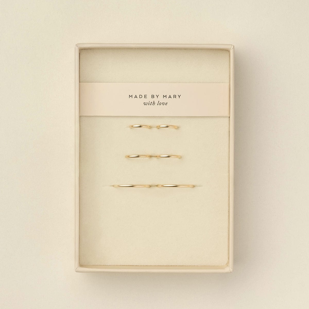 Six delicate gold wire  hoop earrings displayed inside a cream-colored presentation box with a paper band reading 'MADE BY MARY with love'.