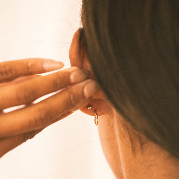 The back of a person's ear, showing a gold round ball earring backing and a thin gold hoop, shown on a solid white background.