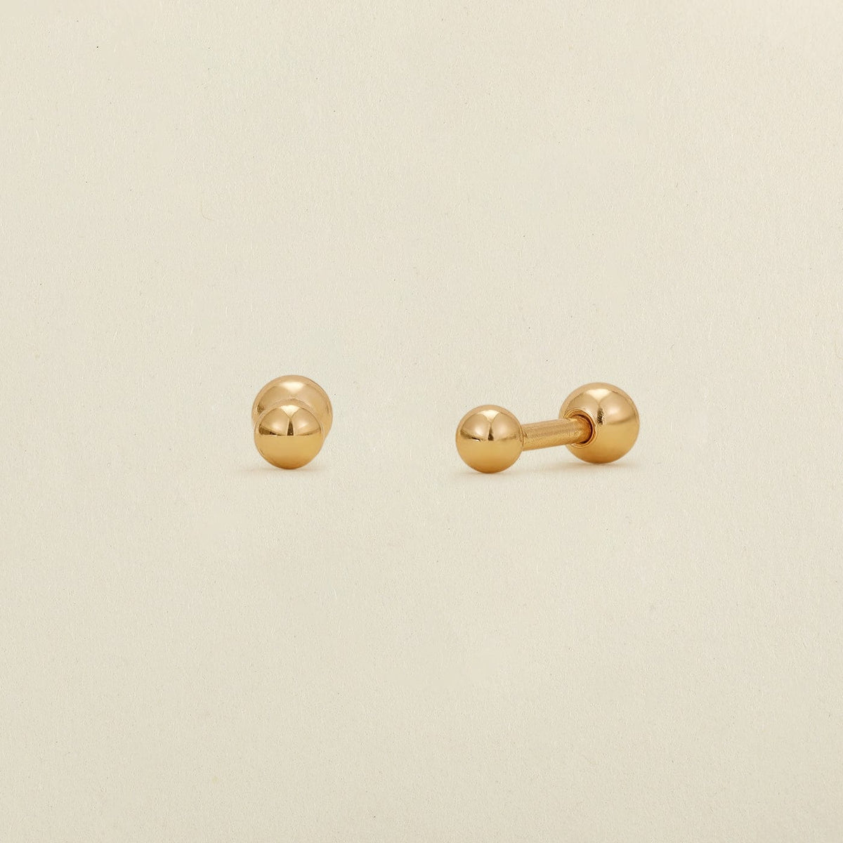 A pair of gold ball stud earrings with round ball backings, shown on a plain, warm off-white background.