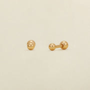 A pair of gold ball stud earrings with round ball backings, shown on a plain, warm off-white background.