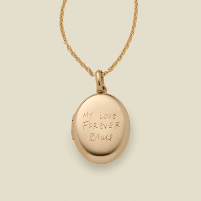 A dainty gold chain necklace with a gold oval locket pendant engraved with handwritten script.