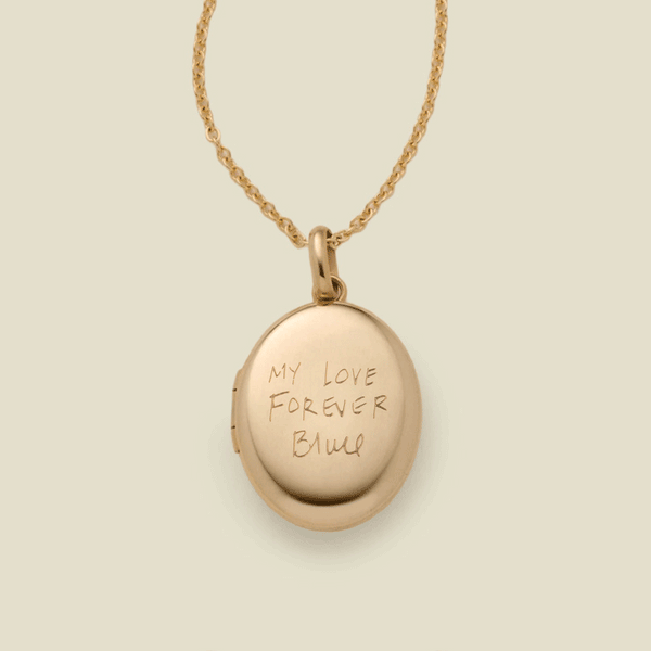 A dainty gold chain necklace with a gold oval locket pendant engraved with handwritten script.