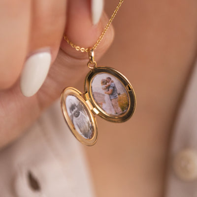 A close-up of a gold oval locket on a delicate gold pendant chain with two photos inside, a black and white photo of a child and a photo of two people embracing.