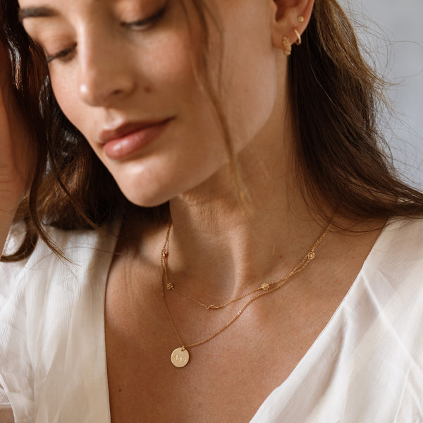 A close-up lifestyle shot of a woman wearing delicate gold layered necklaces, one with a small round disc pendant, and the other with stationary metal birth flowers along the chain, styled with a white top.