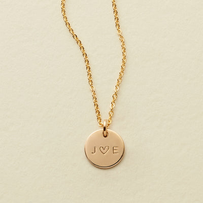 A delicate gold pendant chain necklace with an engraved disc reading 'J ♥ E', shown against a solid light beige background.